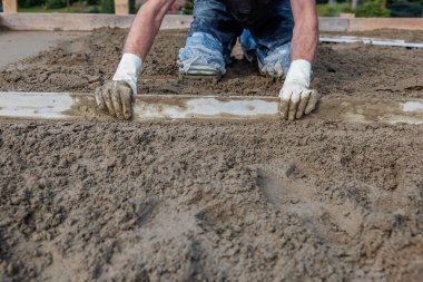 Bir işçi beton zeminde diz çöker, ıslak betonu düzleştirmek için düz bir alet kullanır. Ahşap çerçeve ve ayak izleri devam eden inşaat çalışmalarını gösteriyor..