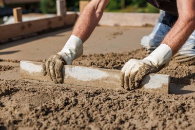 Beyaz eldivenli bir işçinin elleri düz bir aletle ıslak betonu düzleştirir. Tahta şekiller yüzeyi sarar, doğal elementler uzak mesafeden bulanıklaşır..