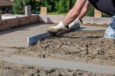 Eldivenli bir işçi, tahta çerçeveli bir yüzeye ıslak beton döşemek için düz bir alet kullanır. Uzakta yeşil bitki örtüsü ve yapılar bulanık görünüyor.