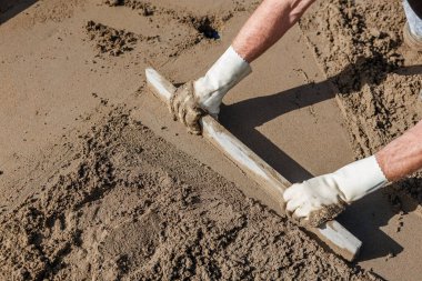 Bir çift eldivenli el ıslak betonu düzleştirmek için düz bir alet kullanır. Yüzey pürüzsüz ve pürüzsüz alanların karışımını gösteriyor, manuel çabaları vurguluyor.