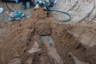 Lastik izleri, el arabası, beton karıştırıcı, hortum ve dağınık alet ve malzemelerle dolu bir kum yığınını gösteren bir inşaat alanı..