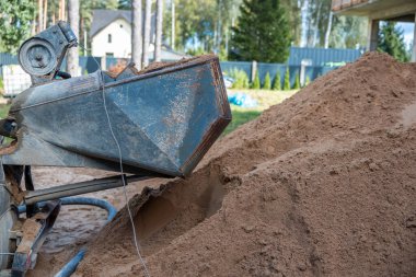 İçinde büyük bir kum yığını, paslı makineler ve çitli bir alan bulunan bir açık hava şantiyesi. Ağaçlar ve bir ev uzaktan görülebilir..