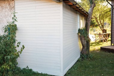 Çimlerle, çalılarla ve sandalyeli ahşap bir masa ile çevrili, eğimli bir ağacın yanında küçük, beyaz bir ahşap kulübe ve panjurlar duruyor..