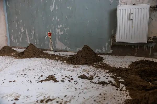 Beyaz zeminde toprak yığınları olan kapalı bir inşaat sahnesi kısmen boyanmış mavi bir duvar, beyaz bir radyatör ve solunda kırmızı vanası olan bir boru..