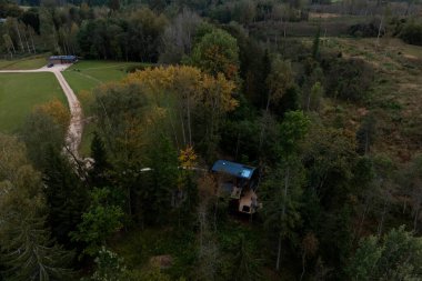 A small modern cabin with a blue roof sits in a forested area with autumn trees, a dirt path, a grassy area, a parked vehicle, and a small structure.