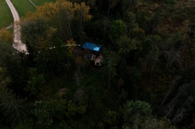 Mavi çatılı küçük bir kulübe, yeşil ve sonbahar renklerinde uzun ağaçlarla çevrili bir orman bölgesinde bulunur. Toprak bir patika açık bir çimenlik alana götürür..