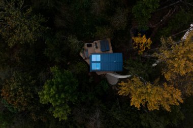 An aerial view of a blue roofed structure with a wooden deck, outdoor furniture, and a curved pathway, surrounded by green and yellow forest foliage.