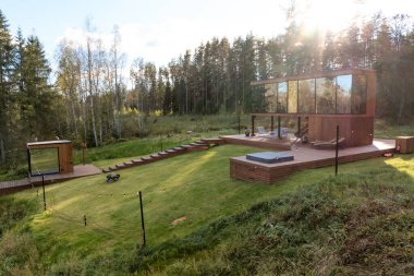 Geniş camlı, modern iki katlı ahşap bir ev. Oturma ve jakuzili bir güverte. Uzun ağaçlarla çevrili..