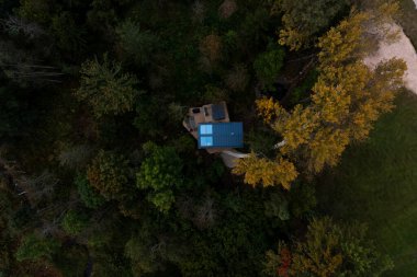 Havadan bakıldığında yeşil ve altın ağaçlardan oluşan bir ormanın içinde mavi bir çatı yapısı görülüyor. Sağ üst köşede açık renkli bir toprak açıklığı görünüyor..