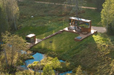 Ahşap bir güvertede büyük cam pencereli modern bir ev, çitlerle çevrili bir alan, yakınlarda küçük bir yapı ve ağaçlar ve bitki örtüsüyle çevrili bir dere..