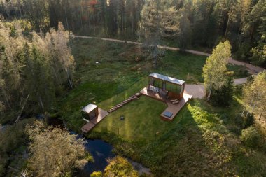 Geniş cam pencereli, ahşap güverteli ve açık hava koltuklu modern bir kulübe. Daha küçük bir birim, ahşap patika ve akarsu ağaçlarla çevrilidir..