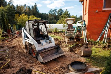 Kirli yolda, kırmızı tuğlalı bir binanın yanında, ağaçlar, bitki örtüsü ve görülebilir beyaz bir sera bulunan beyaz bir fren motoru ve yeşil bir mini kazıcı bulunur..