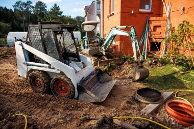 Beyaz fren dümeni yükleyicisi, yeşil kazıcı, merdivenli kırmızı tuğla bina ve seranın yakınındaki dağınık malzemelerin yer aldığı bir inşaat alanı..