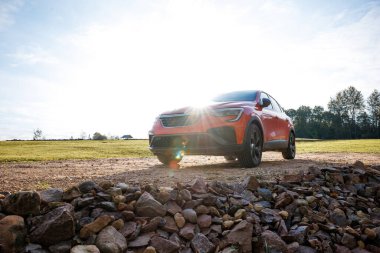 Turuncu bir SUV parlak güneş ışığı altında kirli bir patikada park halinde. Ön planda engebeli kayalar, çimenli tarlalar ve uzaktaki ağaçlar görülebilir..
