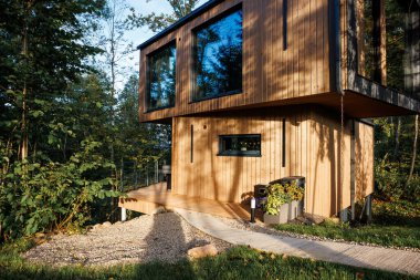 Ağaçlarla çevrili, platformunda büyük cam pencereleri olan modern bir ahşap ev. Yakınlarda bir saksı olan bir çakıl yolu girişe çıkıyor..
