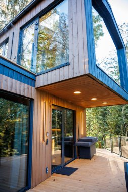 Büyük camlı pencereli iki katlı ahşap bir ev, cam parmaklıklı bir balkon ve uzun ağaçlarla çevrili ahşap bir güverte..