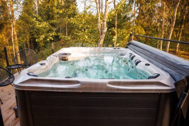 Outdoor hot tub with jet nozzles on a wooden deck, surrounded by tall trees with green and yellow leaves. Black metal chairs and table are nearby.