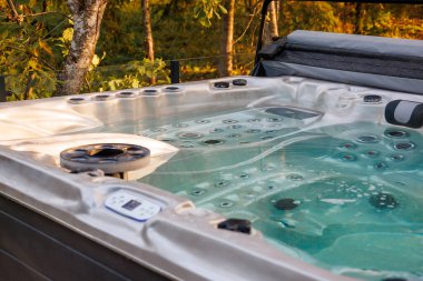 An outdoor hot tub with clear water and active jets, partially covered, surrounded by trees with green and yellow leaves in a private setting.