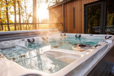 A steaming hot tub with jets sits outdoors by a wooden structure with a glass door. Sunlight filters through tall trees, reflecting on the water.