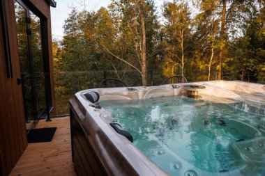 Sonbahar ağaçlarıyla çevrili ahşap bir güvertede kaynayan suyla modern jakuzi. Cam korkuluklar ve siyah sandalyeler orman ortamını güçlendirir.