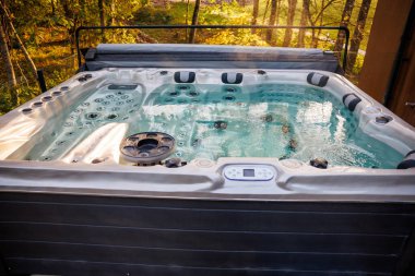 Modern hot tub with ergonomic seating and steaming water on a raised deck, surrounded by tall trees with golden and green autumn leaves.