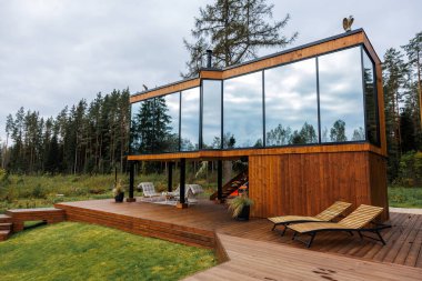 Açısal mimarisi ve büyük cam pencereleri olan modern ahşap bir ev, bulutlu bir gökyüzünün altındaki ağaçlarla çevrili camların üzerinde yükselir..