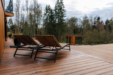 Siyah metal çerçeveli üç tahta şezlong geniş bir ahşap güvertede oturur. Çağdaş bir yapı ve huş ağaçları görülebilir..