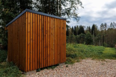 A small wooden structure with vertical planks and a slanted roof stands on gravel, surrounded by grass and trees. String lights are visible in the distance.