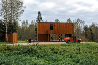 Dikdörtgen ahşap bir ev, ayaklıklar üzerinde minimalist tasarım, yakınlarda turuncu bir araba, ağaçlar ve bulutlu bir gökyüzünün altında çimlerle çevrili..