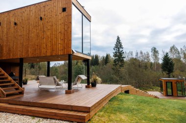 Siyah destekli modern ahşap bir ev büyük cam pencereli geniş bir güverte ve yakınlarda ağaçlar ve otlarla çevrili ayrılmış bir yapı..