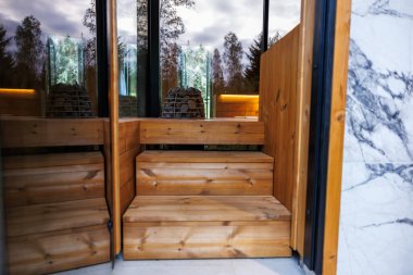 Küçük ahşap bir merdiven, ağaçları yansıtan cam bir kapıya götürür. Mermer bir duvar kontrastı ekler, içinde odun görünür, dışında uzun ağaçlar..