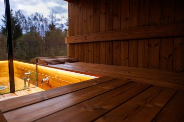 Ilık ışıklandırmalı ahşap sauna, düz banklar, cam panel ağaçları ve bulutlu gökyüzünü gösteren, ve duş alanı olan görünür musluklar..