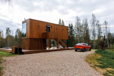 Büyük cam pencereli dikdörtgen ahşap bir ev, dış merdiveni olan camların üzerinde duruyor. Yakınlardaki çakıl yolda kırmızı bir araba var..