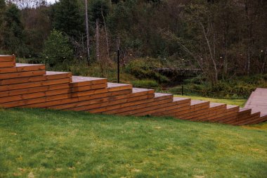 Diagonal wooden steps lead to a flat platform on a grassy slope, with a small bridge and string lights suspended above in a natural setting.