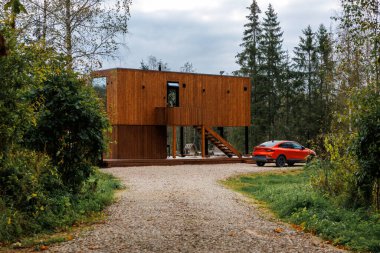 Büyük pencereleri ve dış merdivenleri olan yüksek ahşap bina, çakıl yola kurulmuş. Turuncu bir araba yakınlarda park halinde, ağaçlar ve her yer yeşillikle çevrili..