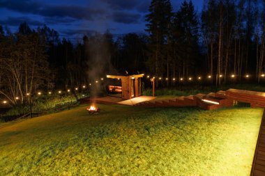 Sıcak ışıklandırması olan ahşap bir yapının gece görüşü, çimenli çimlere duman saçan bir ateş çukuru, ve uzun ağaçların yanındaki ip ışıkları.