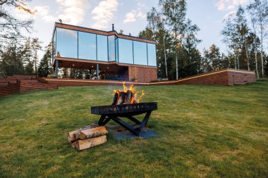 Ahşap dış görünüşlü cam kaplı bir ev ağaçlarla çevrili, çimenli bir yamaçta durur. Ön planda yanan odunları ve yakacak odunları olan bir ateş çukuru var..