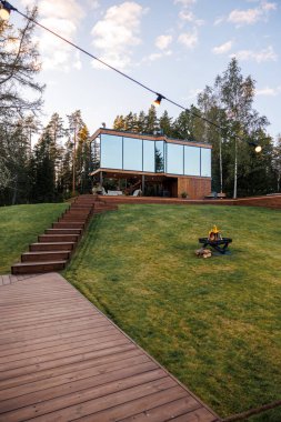 Ahşap dış görünüşlü cam kaplı bir ev ağaçlarla çevrili çimenli bir tepenin üzerinde oturur. Bir ateş çukuru, sicim ışıkları ve ahşap patika sahneyi tamamlar..