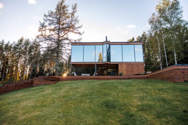Geniş camlı pencereleri ve eğimli çimenli bir alanda ahşap panelleri olan, uzun ağaçlar ve güneş ışığı yansımalarıyla çevrili, modern açılı bir ev..
