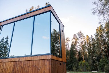 Geniş camlı pencereli modern bir ahşap yapı orman ortamında, yemyeşil ve yaprak döken türler de dahil olmak üzere uzun ağaçların arasında duruyor..