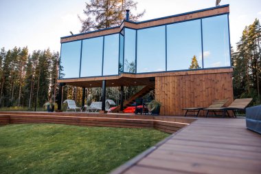 Yüksek ağaçlarla çevrili camlı camlı camlı camlı, iki katlı ahşap bir ev. Açık havada oturma, merdiven ve ahşap bir güverte..