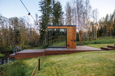 Çimenler, ağaçlar ve metal bir merdiveni olan küçük bir su kütlesiyle çevrili, yükseltilmiş bir güvertede büyük camdan penceresi olan modern bir ahşap kulübe..