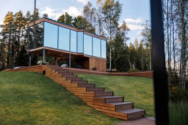 Camdan duvarları olan modern bir ev ayaklıklar tarafından desteklenen çimenli bir yamaçta duruyor. Yansıtıcı pencereler ağaçların yakınındaki, tahta merdivenli aynalar..