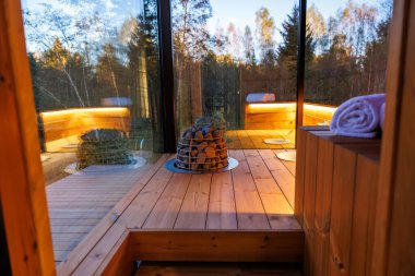 İçinde cam duvarlar bulunan modern ahşap bir saunanın içi, taşlı merkezi ısıtıcı, ahşap banklar, sıcak ışıklandırma, ve yan tarafında katlanmış bir havlu..