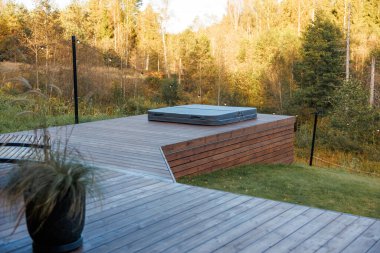 Outdoor wooden deck with a covered hot tub, bordered by grass and autumn trees. String lights and a potted plant add decorative elements.