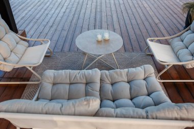 Cushioned chairs and sofa surround a round table with candles on a patterned rug atop a wooden deck with clean, expansive wooden planks.