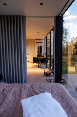 Camdan duvarları olan modern bir yaşam alanı, örtülü bir yatak, siyah sandalyeli bir yemek alanı ve dışarıdaki ağaçların manzarası..