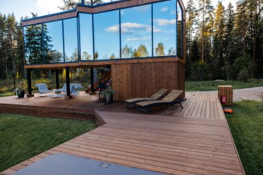 Geniş yansıtıcı pencereli modern ahşap bir ev şezlonglu yüksek bir güverte ve güvertenin kenarında görünür bir yangın söndürücü..