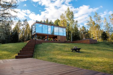 Çimen ve ağaçlarla çevrili, ahşap platformda yükseltilmiş modern cam duvarlı bina. Yansıtıcı pencereler, ahşap merdivenler ve çimenlikte görünen ateş çukuru..