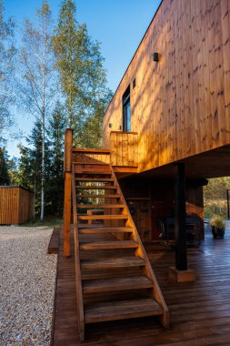 Minimalist ahşap bir kulübe, dış merdiveni olan, uzun huş ağaçları ve yemyeşil ile çevrili, yakındaki çakılları olan ahşap bir güvertede dinleniyor..